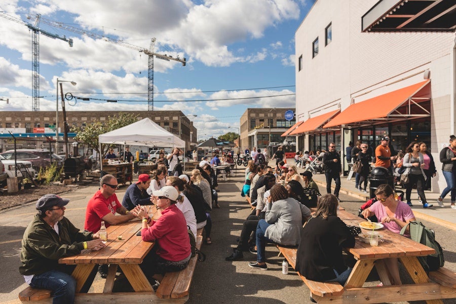 Groups of people enjoying food and drinks at outdoor picnic tables on a sunny day.
