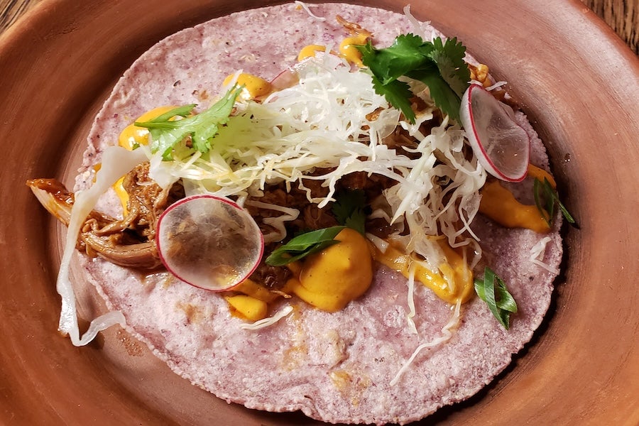 A taco served on a purple corn tortilla, topped with shredded meat, cabbage, radish slices, and fresh cilantro, with drizzles of a creamy orange sauce. The dish is presented on a rustic clay plate.