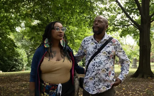 Two people in a leafy park, one holding a phone, both wearing patterned outfits.

